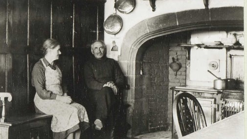 A black and white photograph of Mr and Mrs Lilburn sat in front of a large fireplace at Lindisfarne Castle with a dog walking in front of them